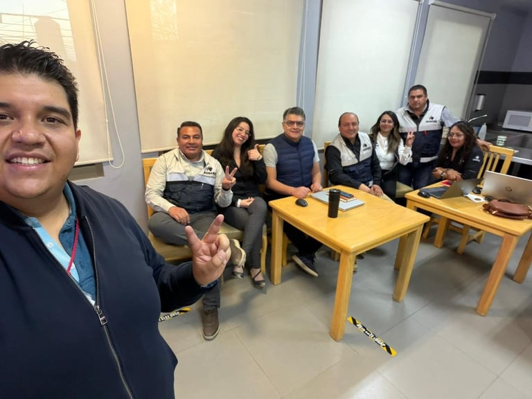 A man in the foreground takes a selfie with a group of coworkers seated at wooden tables in a casual office setting